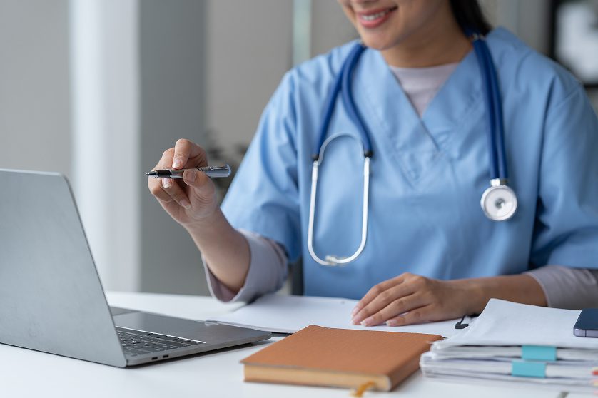 A smiling female medical professional in blue scrubs and a stethoscope uses a laptop and holds a pen at a desk with documents and a notebook.